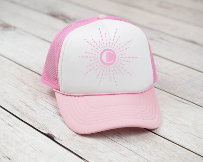 Pink and white trucker hat with single letter monogram set in a sunburst pattern. 