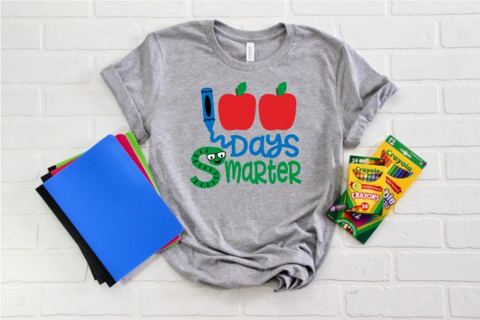 Grey T-shirt with some school supplies staged around it and the 100 Days smarter SVG file cut and applied in iron on.