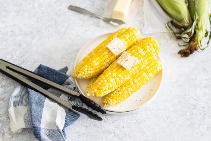 Corn on the cob with butter and pepper on plates with a pair of tongs and a dish towel