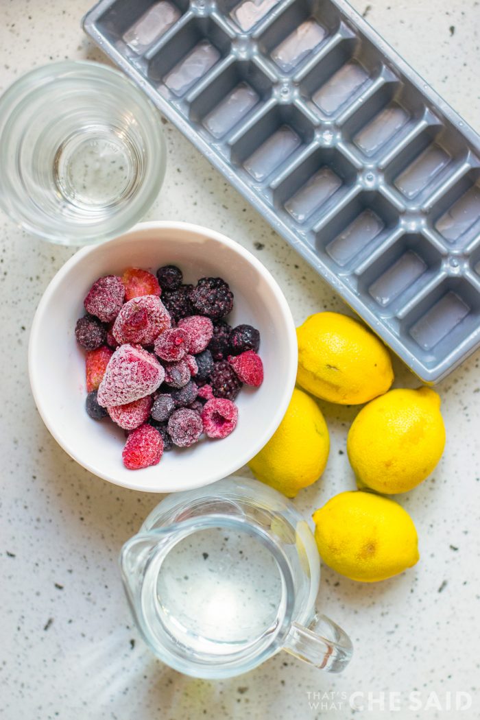 Table top overhead view of pitcher with water, bowl of frozen berries, lemons, cups and ice cube tray.