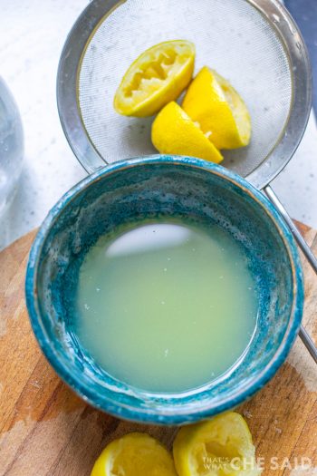 Small blue bowl filled with lemon juice and a strainer on the side with juiced lemons inside.