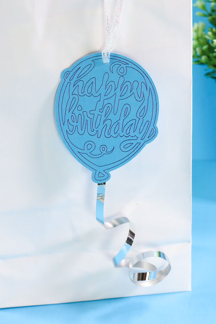 Happy Birthday Balloon Gift tag with ribbon tail hanging from a white gift bag