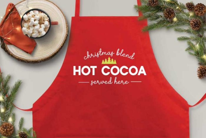 Red Apron with "Christmas Blend Hot Cocoa Served Here" with a mug of hot cocoa and some christmas greens as accents. Horizontal format