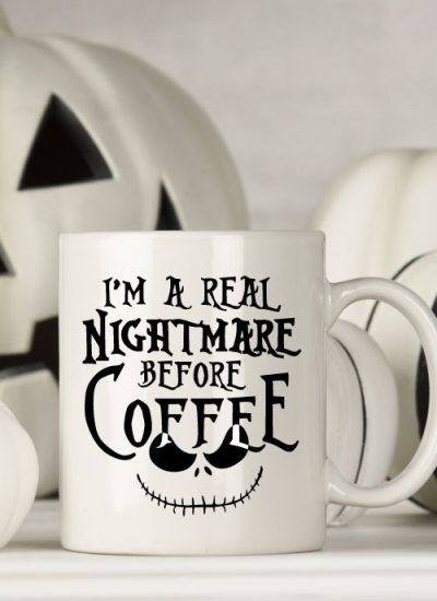 White mug with black and white halloween decor behind. Mug Reads" I'm a Real Nightmare Before Coffee"