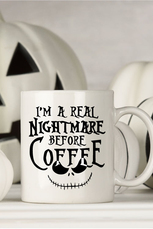 White mug with black and white halloween decor behind. Mug Reads" I'm a Real Nightmare Before Coffee"