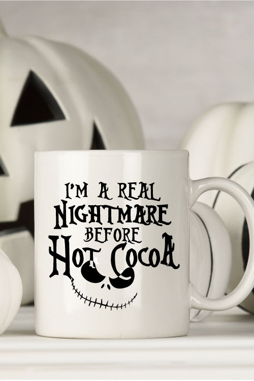 White mug with black and white halloween decor behind. Mug Reads" I'm a Real Nightmare Before Hot Cocoa"