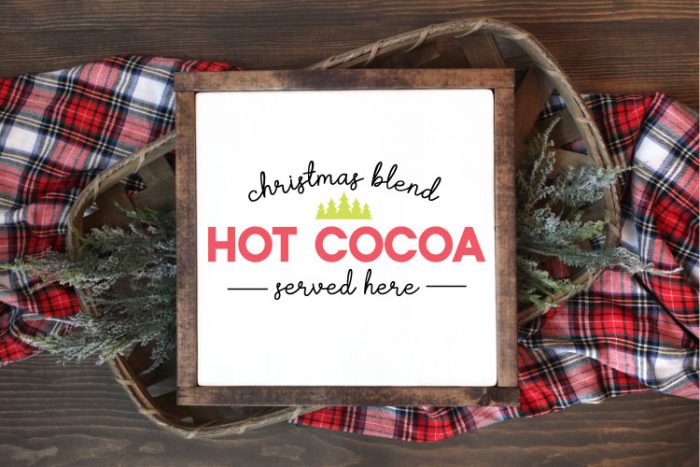 Square sign with white background and wooden frame laying one a basket of evergreens snips and a plaid fabric. "Christmas Blend Hot Cocoa Served here" is applied in vinyl