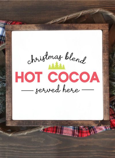 Square sign with white background and wooden frame laying one a basket of evergreens snips and a plaid fabric. "Christmas Blend Hot Cocoa Served here" is applied in vinyl - vertical