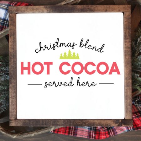 Square sign with white background and wooden frame laying one a basket of evergreens snips and a plaid fabric. "Christmas Blend Hot Cocoa Served here" is applied in vinyl - vertical