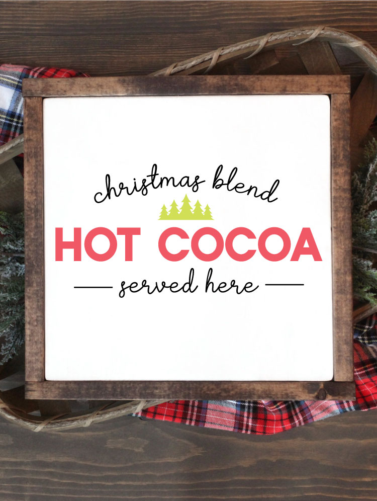 Square sign with white background and wooden frame laying one a basket of evergreens snips and a plaid fabric. "Christmas Blend Hot Cocoa Served here" is applied in vinyl - vertical