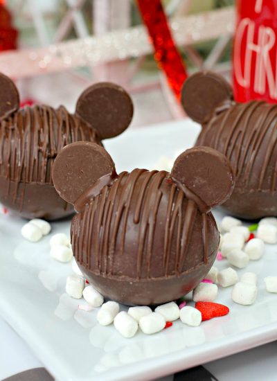 Three Chocolate Hot Chocolate Bomb with added chocolate wafers as ears to resemble Mickey Mouse with marshmallows and Christmas decor in background vertical orientation 45 degrees angle