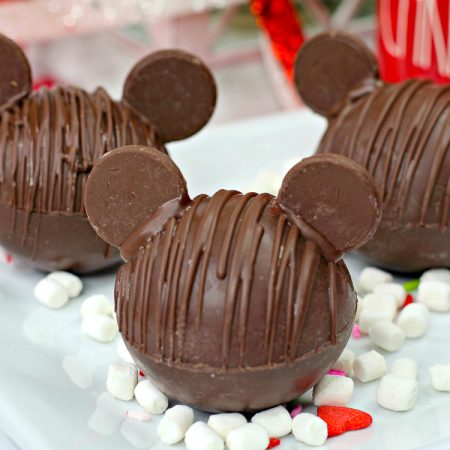 Three Chocolate Hot Chocolate Bomb with added chocolate wafers as ears to resemble Mickey Mouse with marshmallows and Christmas decor in background vertical orientation 45 degrees angle