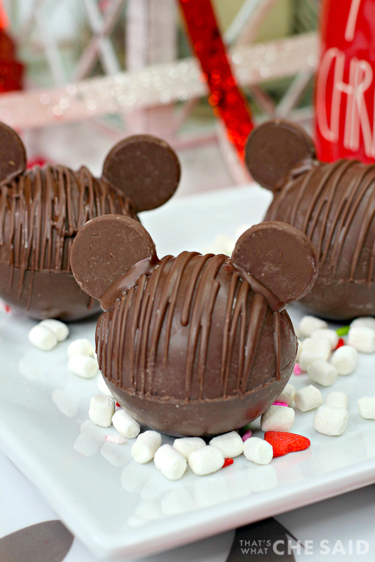Three Chocolate Hot Chocolate Bomb with added chocolate wafers as ears to resemble Mickey Mouse with marshmallows and Christmas decor in background vertical orientation 45 degrees angle