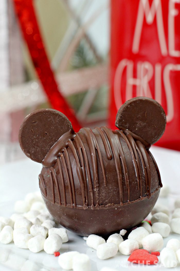 Chocolate Hot Chocolate Bomb with added chocolate wafers as ears to resemble Mickey Mouse with marshmallows and Christmas decor in background vertical orientation