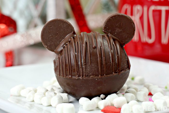 Chocolate Hot Chocolate Bomb with added chocolate wafers as ears to resemble Mickey Mouse with marshmallows and Christmas decor in background horizontal orientation