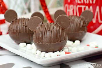 Three Chocolate Hot Chocolate Bomb with added chocolate wafers as ears to resemble Mickey Mouse with marshmallows and Christmas decor in background