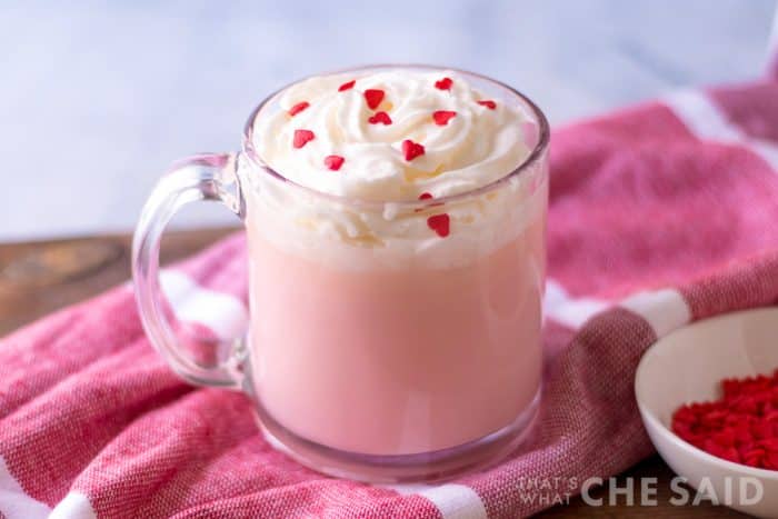 4 ingredient pink hot chocolate in clear mug with whipped topping and heart sprinkles on pink towel horizontal close up