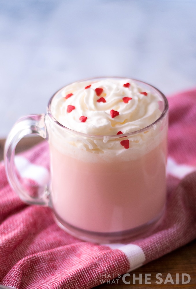 4 ingredient pink hot chocolate in clear mug with whipped topping and heart sprinkles on pink towel close up