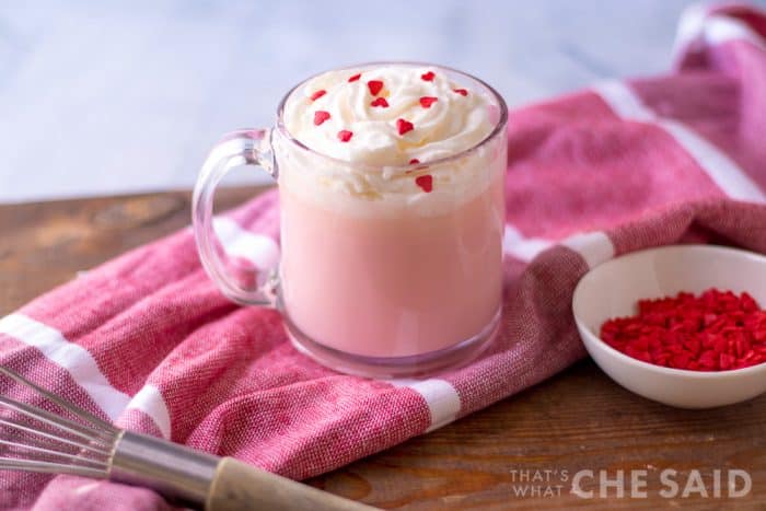 4 ingredient pink hot chocolate in clear mug with whipped topping and heart sprinkles on pink towel horizontal orientation