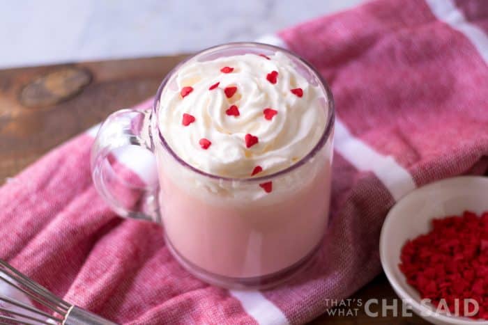 4 ingredient pink hot chocolate in clear mug with whipped topping and heart sprinkles on pink towel top down orientation