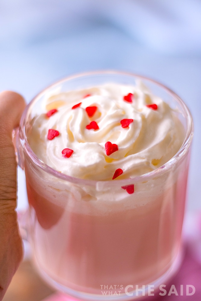 Person holding pink hot chocolate in clear mug with whipped topping and heart sprinkles on pink towel vertical orientation