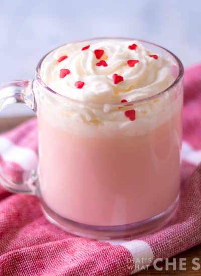 4 ingredient pink hot chocolate in clear mug with whipped topping and heart sprinkles on pink towel square orientation