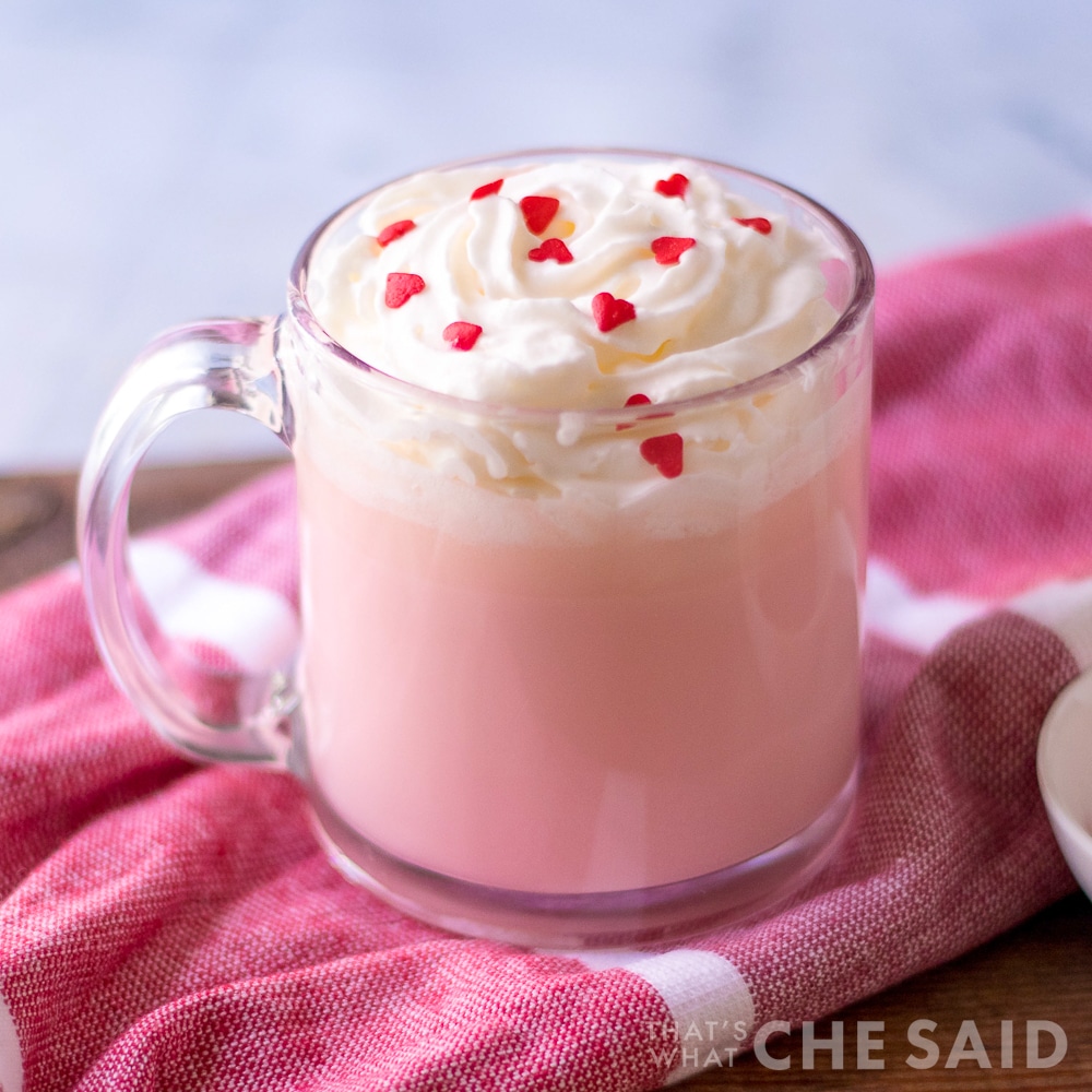 4 ingredient pink hot chocolate in clear mug with whipped topping and heart sprinkles on pink towel square orientation