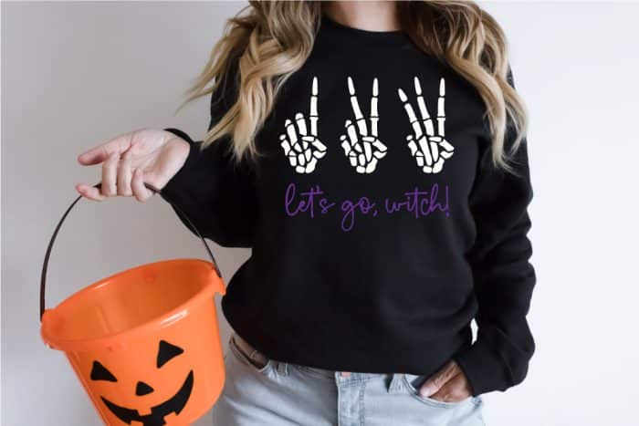 Woman wearing black sweatshirt holding orange pumkin basket with 123 Let's go Witch design on the sweatshirt in horizontal photo format