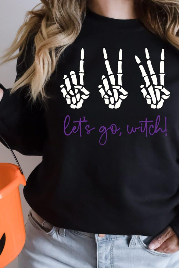 Woman wearing black sweatshirt holding orange pumkin basket with 123 Let's go Witch design on the sweatshirt in vertical photo format