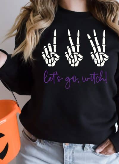 Woman wearing black sweatshirt holding orange pumkin basket with 123 Let's go Witch design on the sweatshirt in square photo format