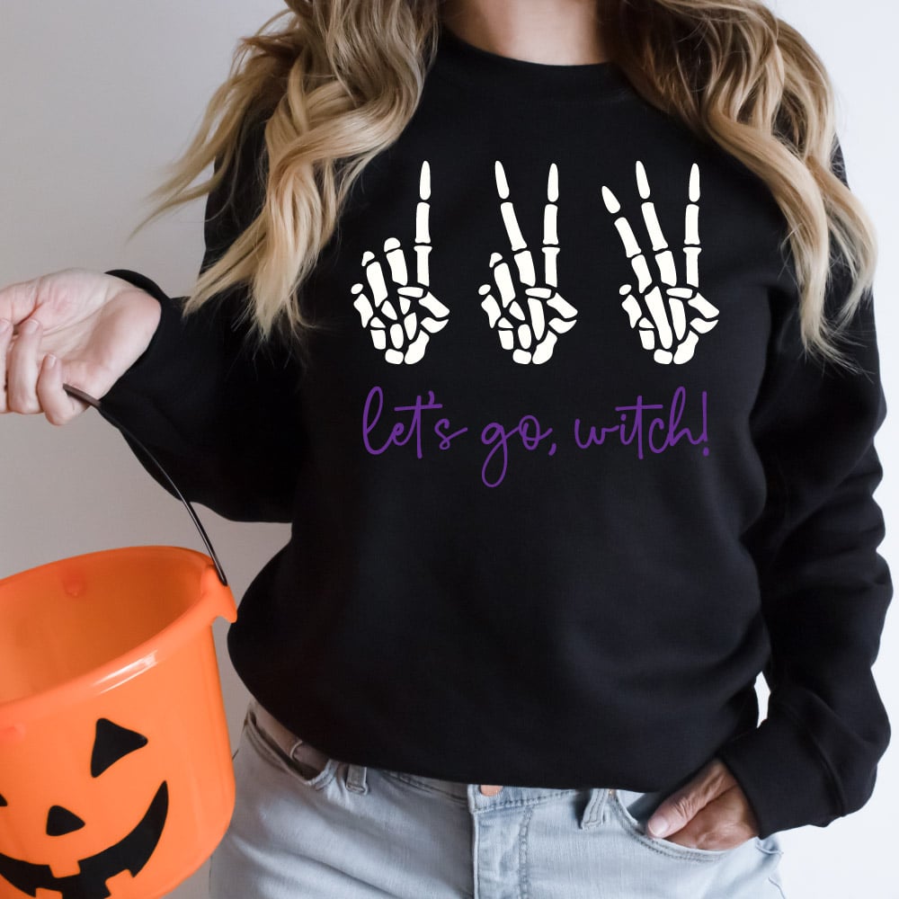 Woman wearing black sweatshirt holding orange pumkin basket with 123 Let's go Witch design on the sweatshirt in square photo format