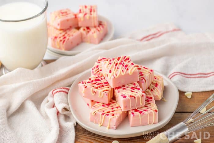 Valentine Sugar Cookie Fudge on a white plate with a towel and a small plate of fudge in background with a glass of milk. - horizontal