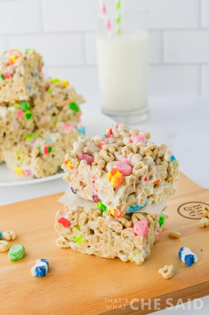Lucky charms treat bars stacked on a cutting board with a plate full and glass of milk in background