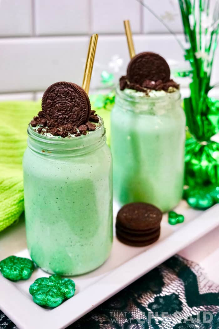 Vertical shot of two oreo mint milkshakes in mason jars with green towel and shamrock decor and gold straws