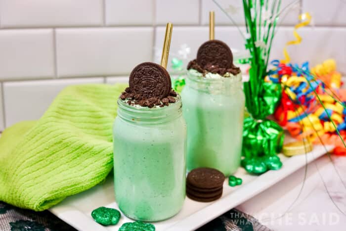 Vertical image of two oreo mint milkshakes in mason jars with green towel, shamrock and rainbow decor