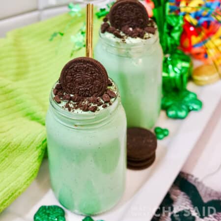 Square image of two oreo mint milkshakes in mason jars with green towel and shamrock decor