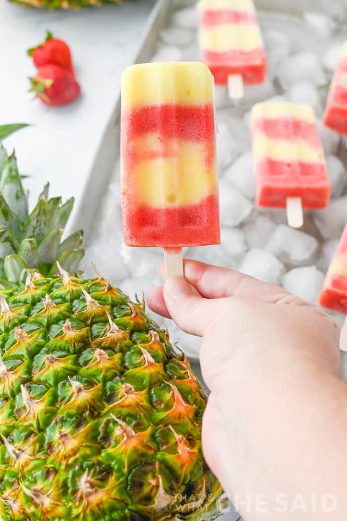 hand holding Lava flow popsicle with pineapple and strawberries in the background