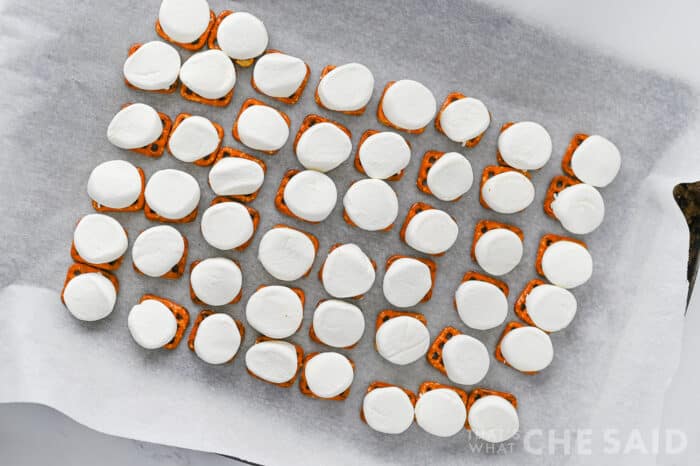 Placing marshmallows on top of square pretzles on a parchment lined sheet pan