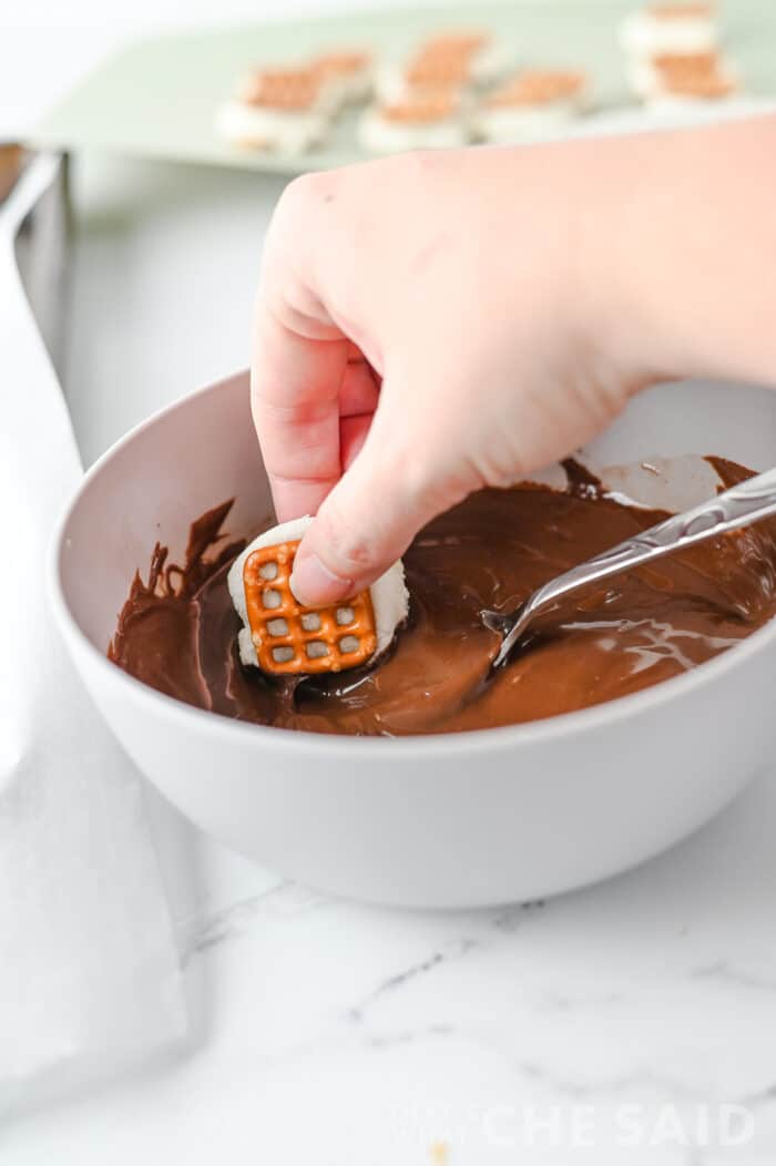 dipping a smore's pretzel bite into melted chocolate in a white bowl